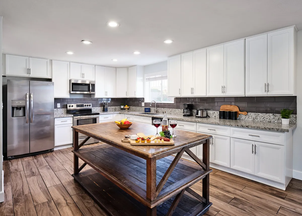 This image showcases a modern kitchen with a central wooden island, granite countertops, stainless steel appliances, and a fruit bowl with wine glasses neatly arranged. The space is stylish yet approachable, inviting guests to cook, entertain, or simply enjoy a meal. Natural light and thoughtful details highlight the kitchen's turnkey amenities.