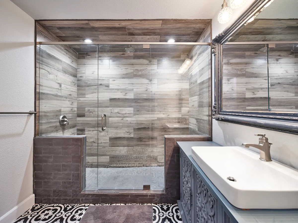 A spa-inspired bathroom with a glass shower enclosure, a stylish vanity with decorative detailing, intricate floor tiles, and a framed mirror. The space exudes serenity and modern luxury—ideal for refreshing showers or self-care routines.