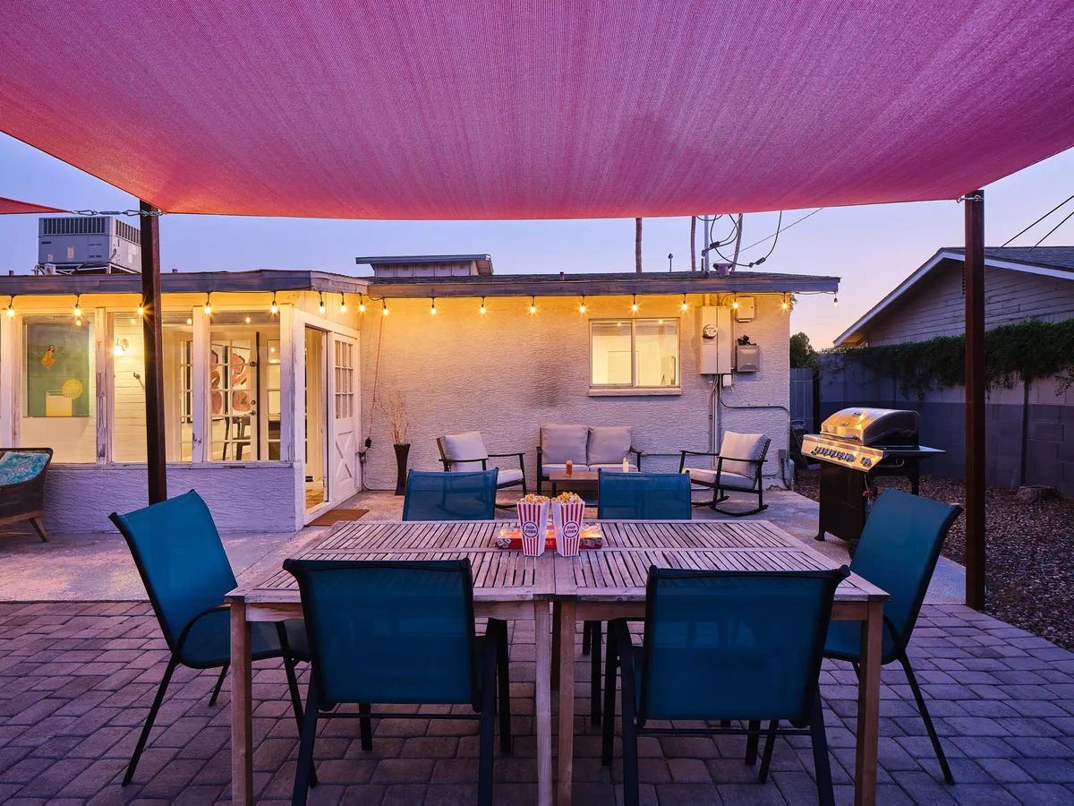 A cheerful backyard oasis featuring a large outdoor dining table beneath string lights, a barbecue grill, and comfortable seating. The inviting space is ideal for outdoor dining, social gatherings, and unwinding after a day of activities.