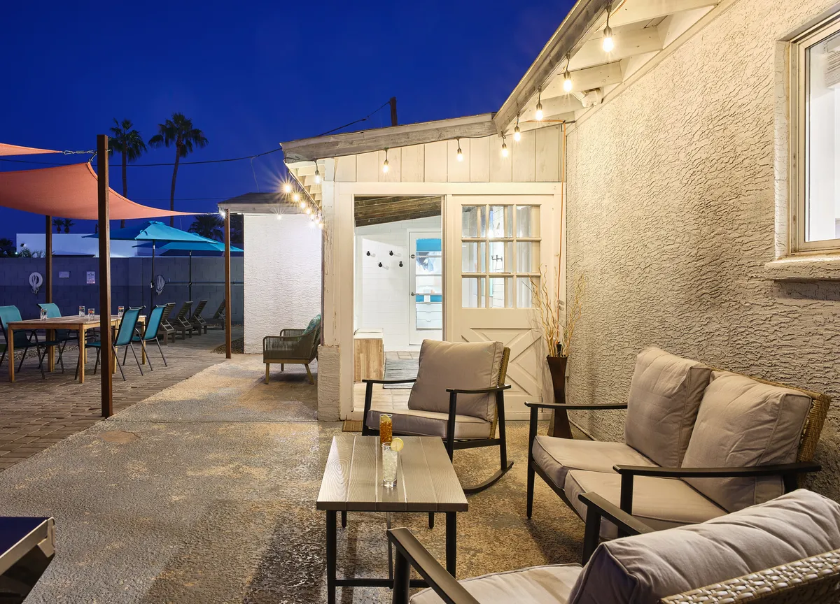 A welcoming outdoor patio scene with string lights, a cozy seating area, and an open door leading inside. The atmosphere is casual and inviting, perfect for groups to relax, socialize, or gather by the fire pit under the stars after a night out.