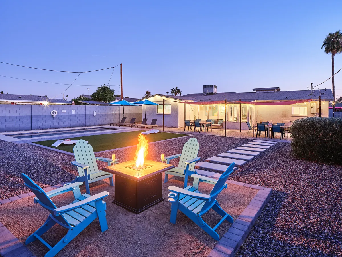 This cozy backyard scene centers on a fire pit surrounded by Adirondack chairs, with the pool, outdoor dining area, and patio visible in the background. The space is set up for communal gatherings, whether stargazing by the fire, socializing, or enjoying sunset moments near the water.