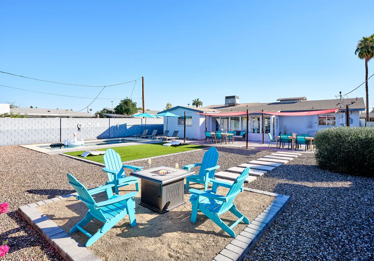 A wide view of the backyard showcases blue Adirondack chairs arranged around a fire pit, a green lawn, a swimming pool, and a spacious patio area with tables. The layout highlights space for swimming, outdoor dining, barbecuing, and socializing, all within a fully enclosed, pet-friendly yard setting.