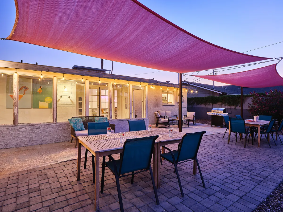 A spacious backyard with outdoor dining tables beneath shade sails, illuminated by string lights, and surrounded by lounge chairs. The setting invites groups to gather for meals, casual meetings, or evening celebrations under a cozy, festive atmosphere.