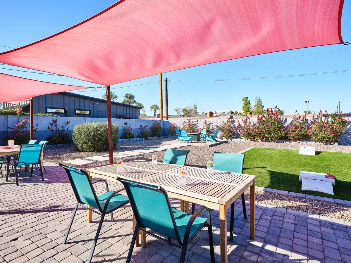 A cheerful backyard scene featuring red shade sails casting cooling shadows over a wooden dining table and blue Adirondack chairs, all surrounded by desert landscaping. The enclosed outdoor space invites relaxation and play, with abundant room for gatherings or pet activities.