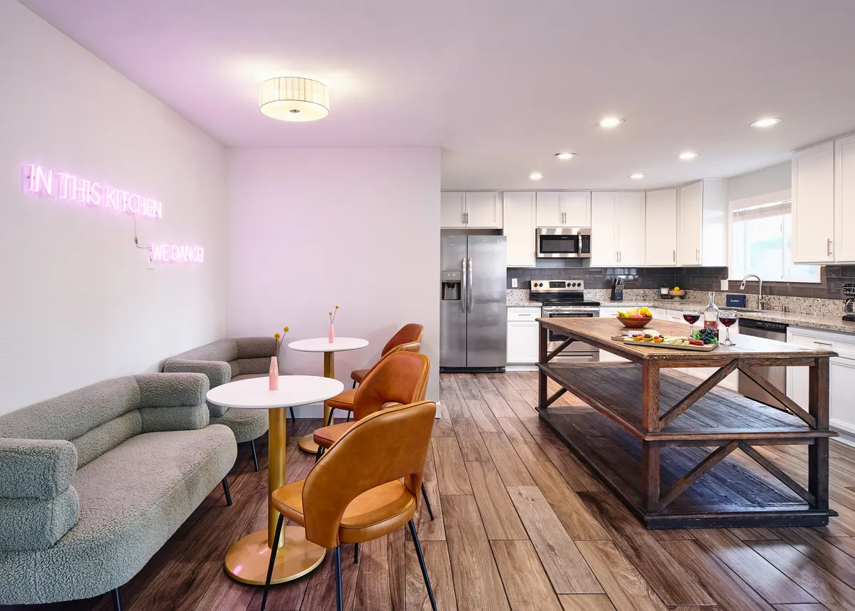 A vibrant kitchen scene with a neon sign reading 'IN THIS KITCHEN WE DINE,' a central kitchen island, and a stylish seating area. The space is designed for both cooking and socializing, radiating a cheerful and inviting energy.