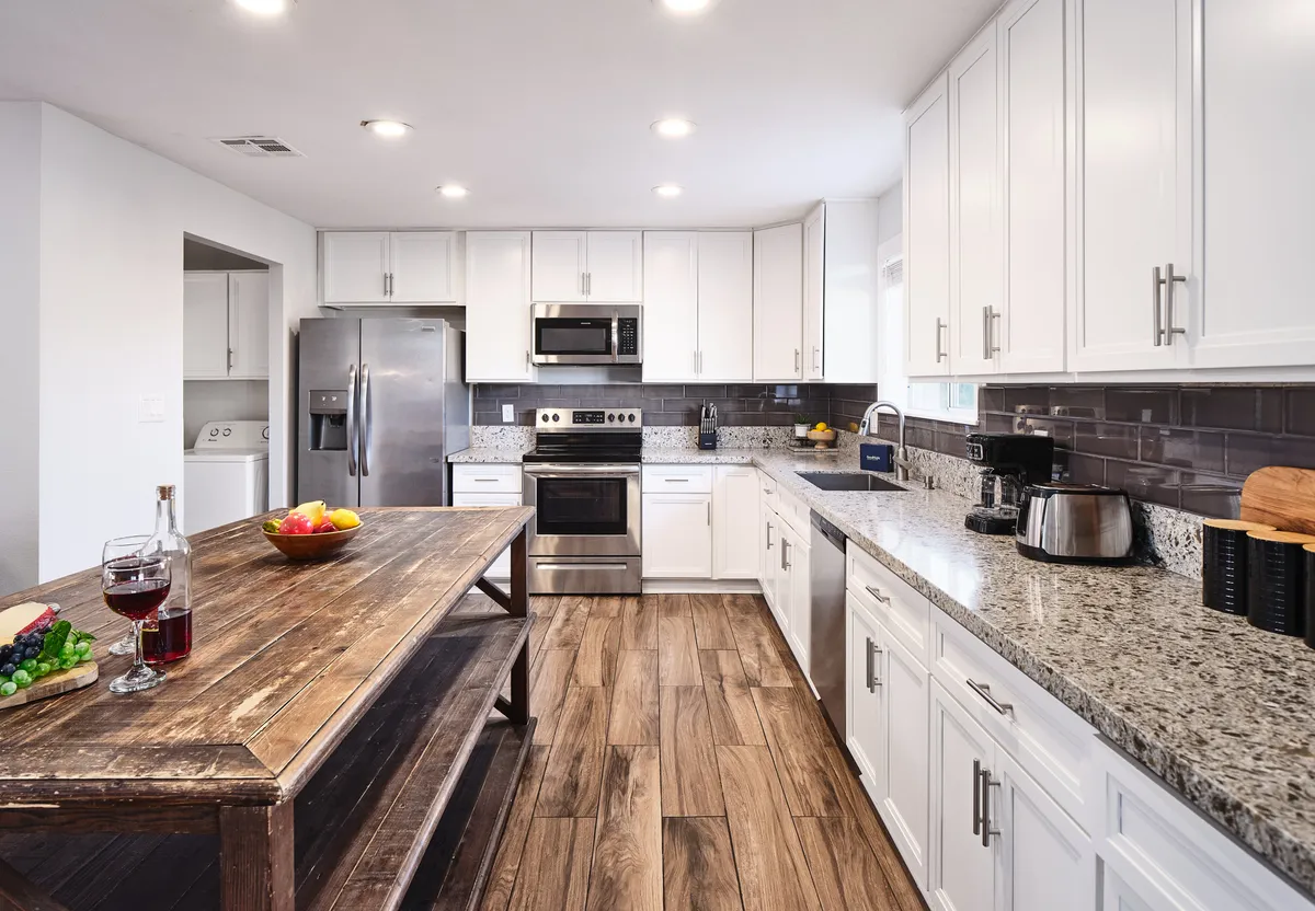 A spacious dining area featuring a large wooden table set for eight, granite countertops, modern stainless steel appliances, a colorful fruit bowl, and wine glasses. The space is warm and inviting, ideal for group gatherings and family meals.